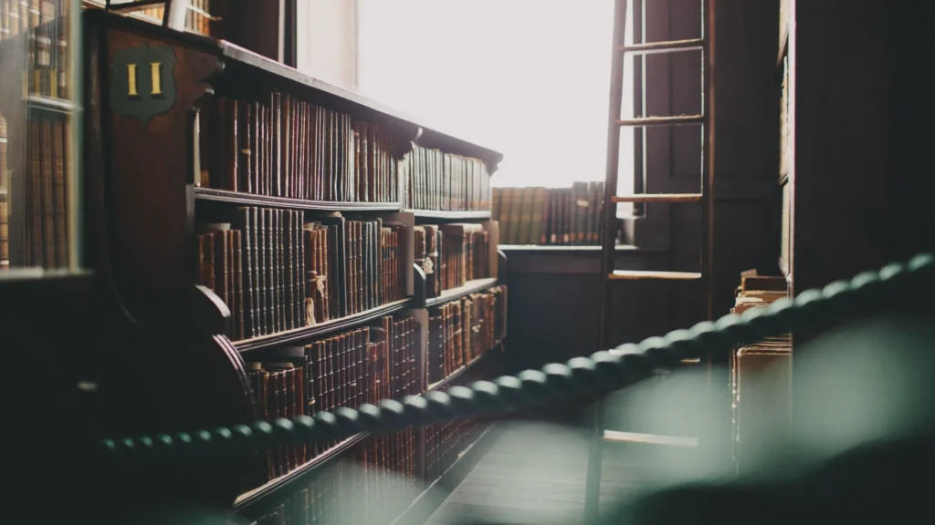 Bookshelves lined with old books, including volumes on the New Testament, a ladder leaning against the wall, and sunlight streaming through a window in a quiet library setting. Advent Hill Ministries