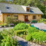 A yellow wooden house with a tiled roof is surrounded by a well-maintained garden, various plants, and green benches—an inviting scene that captures the charm of country living on a sunny day. Advent Hill Ministries
