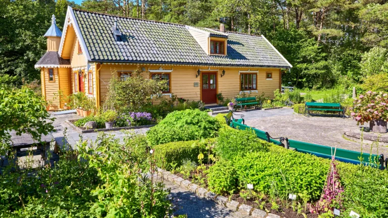 A yellow wooden house with a tiled roof is surrounded by a well-maintained garden, various plants, and green benches—an inviting scene that captures the charm of country living on a sunny day. Advent Hill Ministries